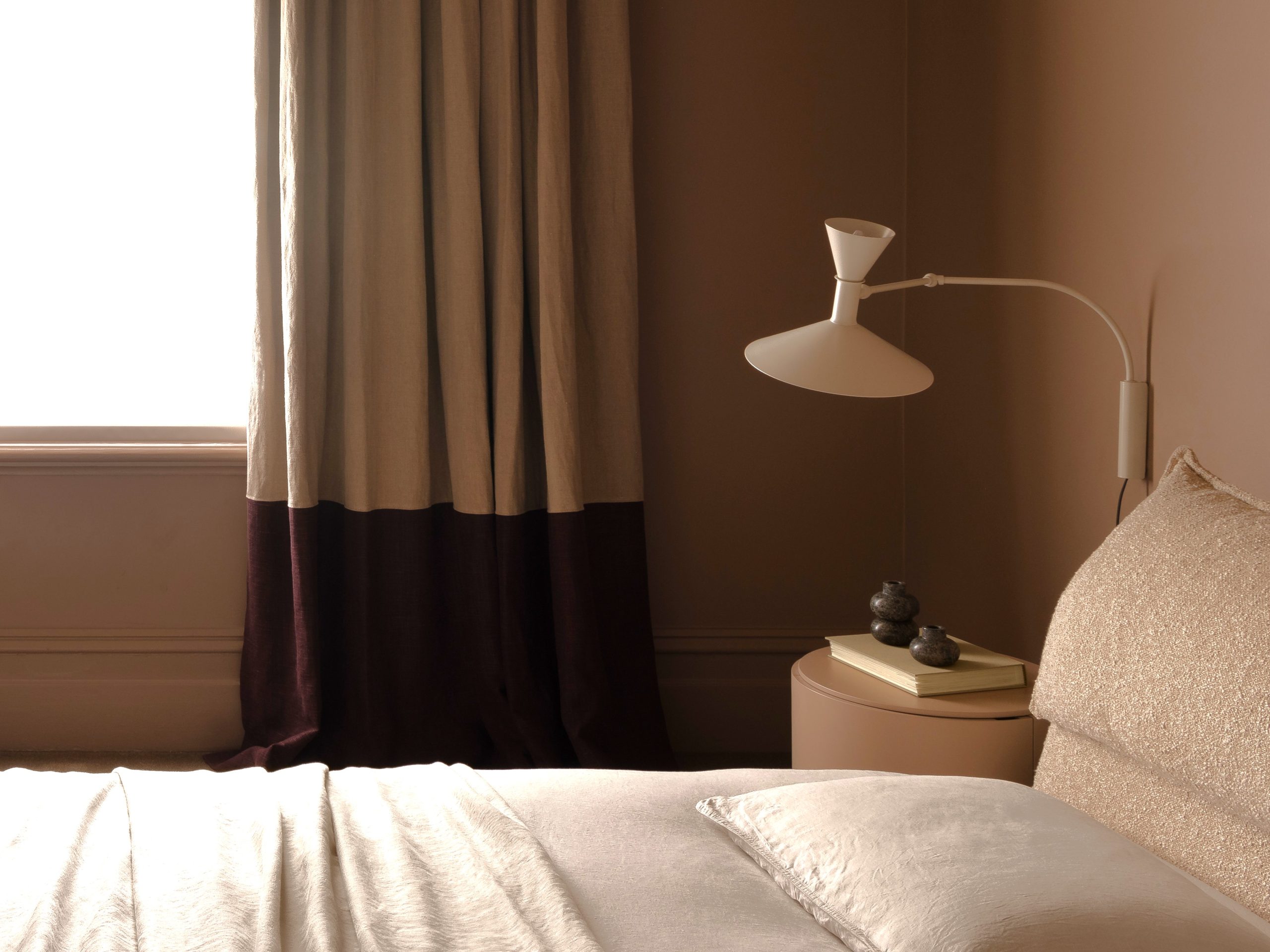 Master bedroom showing lower part of custom window furnishings - banded curtain, bedside table and wall mounted light with book and two small empty vases on display